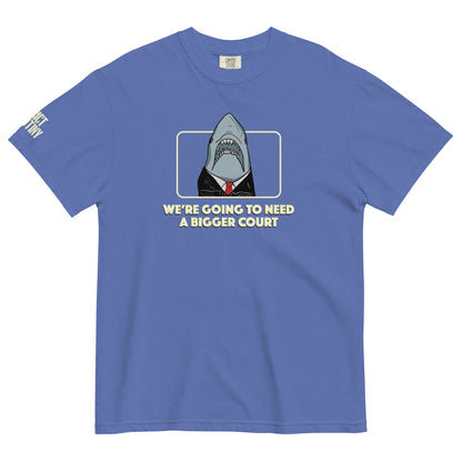 A blue short-sleeve t-shirt with a graphic of a shark dressed in a black suit and red tie, framed by a light yellow border, with the text 'WE'RE GOING TO NEED A BIGGER COURT' printed below. Strict Scrutiny logo in light yellow is visible on the left sleeve